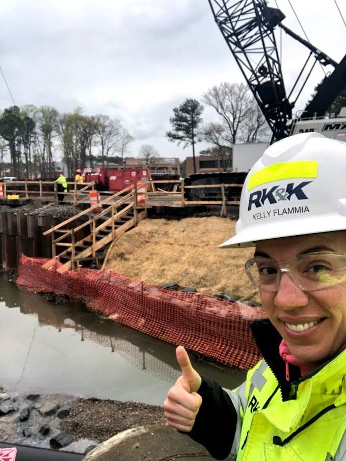Construction site with RK&K worker in safety gear giving a thumbs-up near water and heavy equipment.