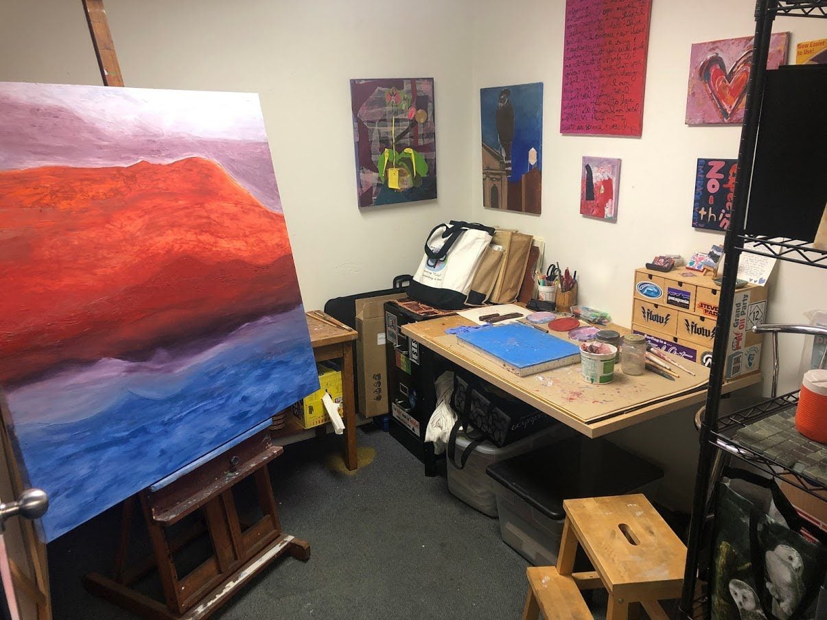Small art studio with a large colorful painting on an easel, a worktable with supplies, and artwork on the walls.