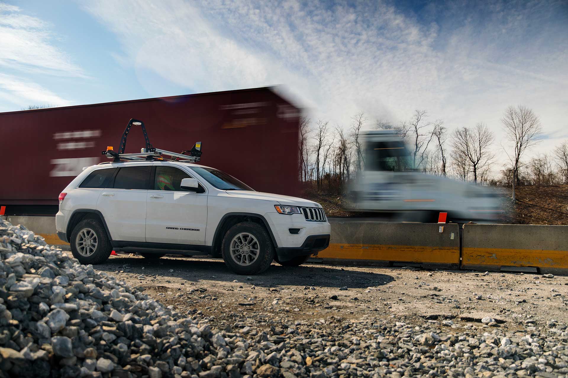 White SUV with roof‑mounted surveying equipment parked on a gravel worksite beside a concrete barrier, with freight vehicles moving past in the background.