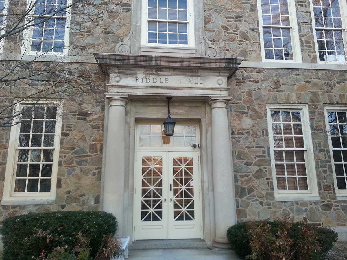 Entrance to Biddle Hall with a stone façade, tall windows, and double doors framed by columns and a hanging lantern.
