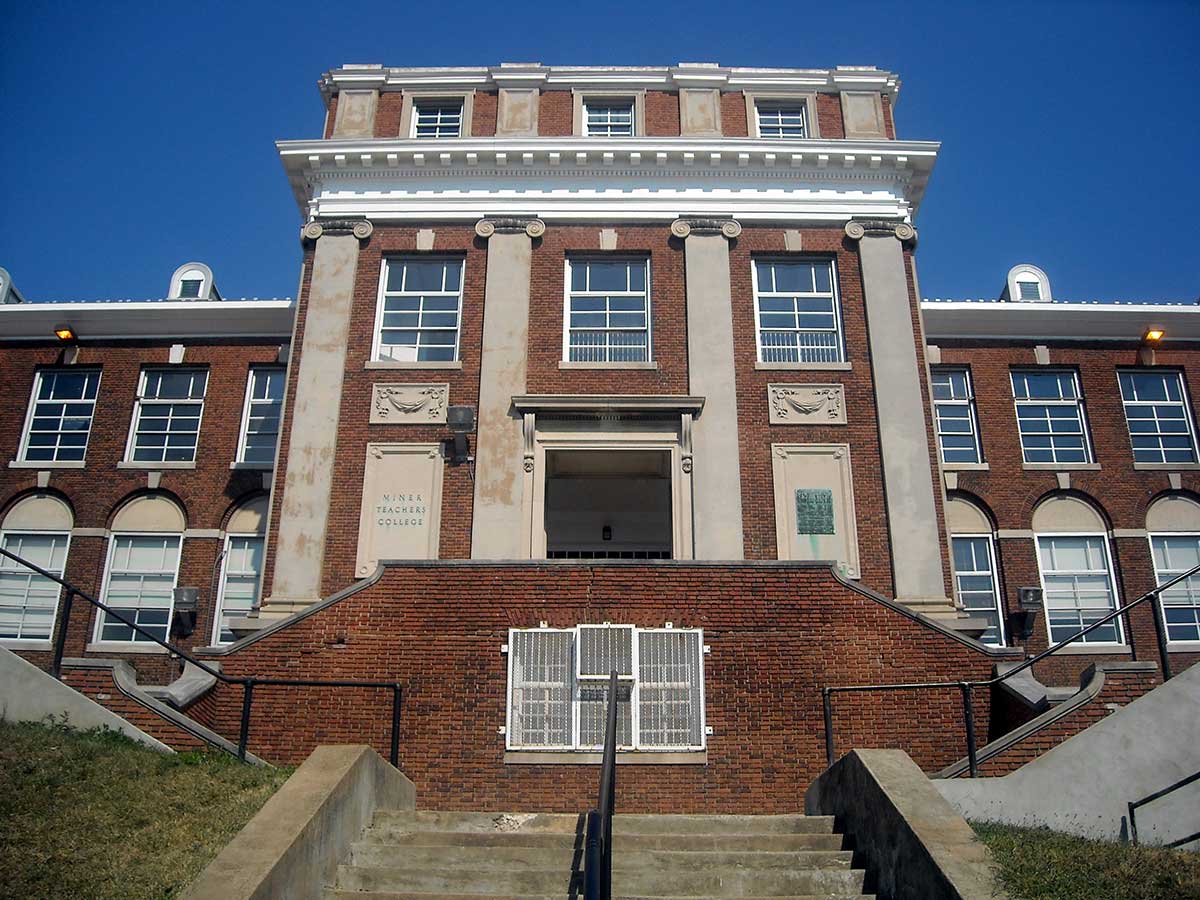 Historic brick building with tall windows, decorative stone details, and a grand central entrance reached by a wide staircase.