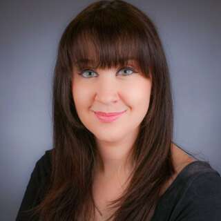 Woman with long, straight dark hair wearing a dark top against a gray studio background.