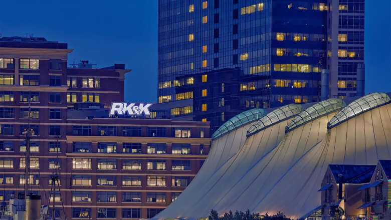 Dusk city skyline with lit office towers, RK\&K sign visible, and a sweeping white curved canopy in the foreground.