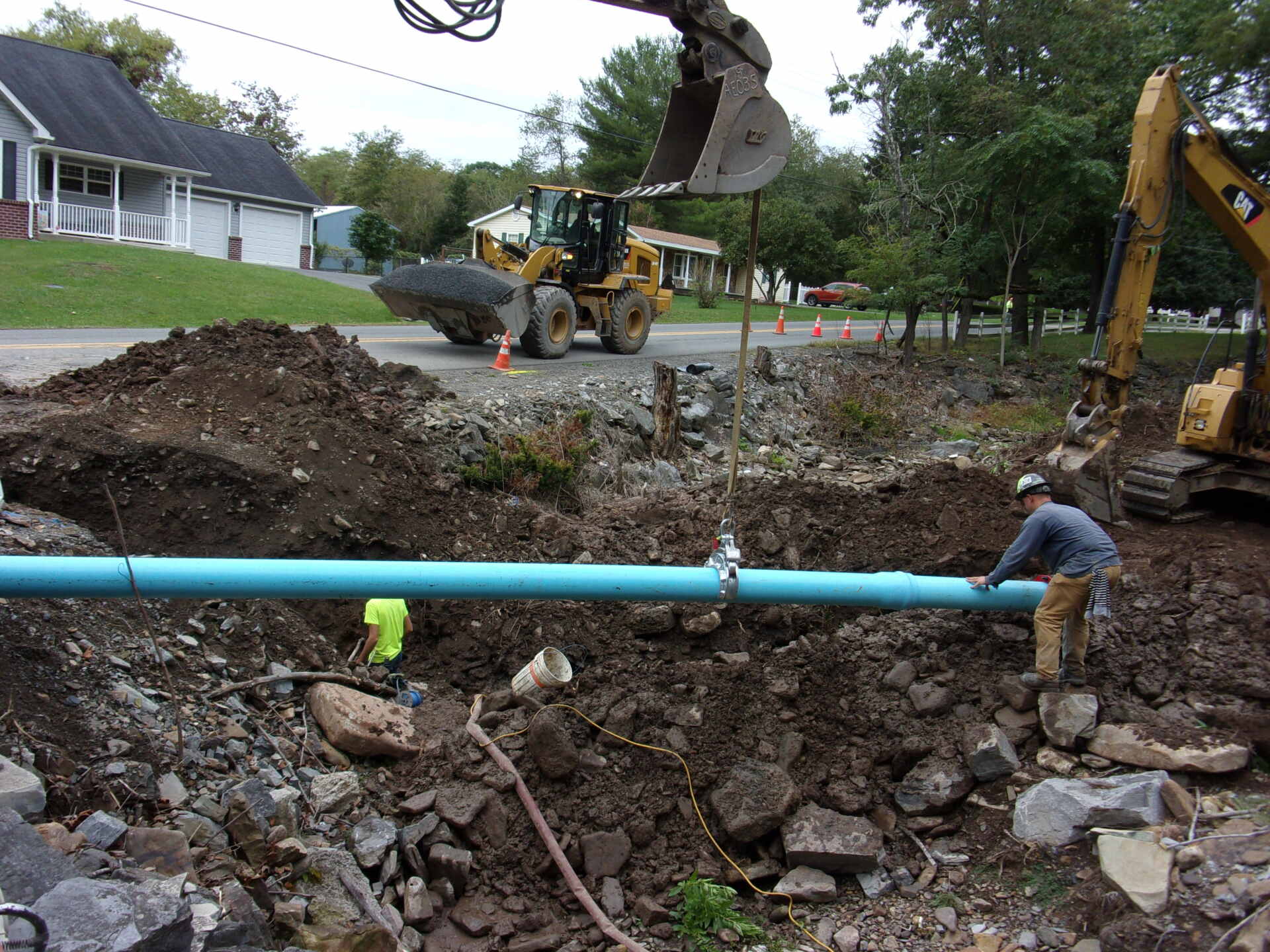 Excavation site with workers and an excavator installing a long blue water pipe in a rocky trench, surrounded by dirt and construction equipment.