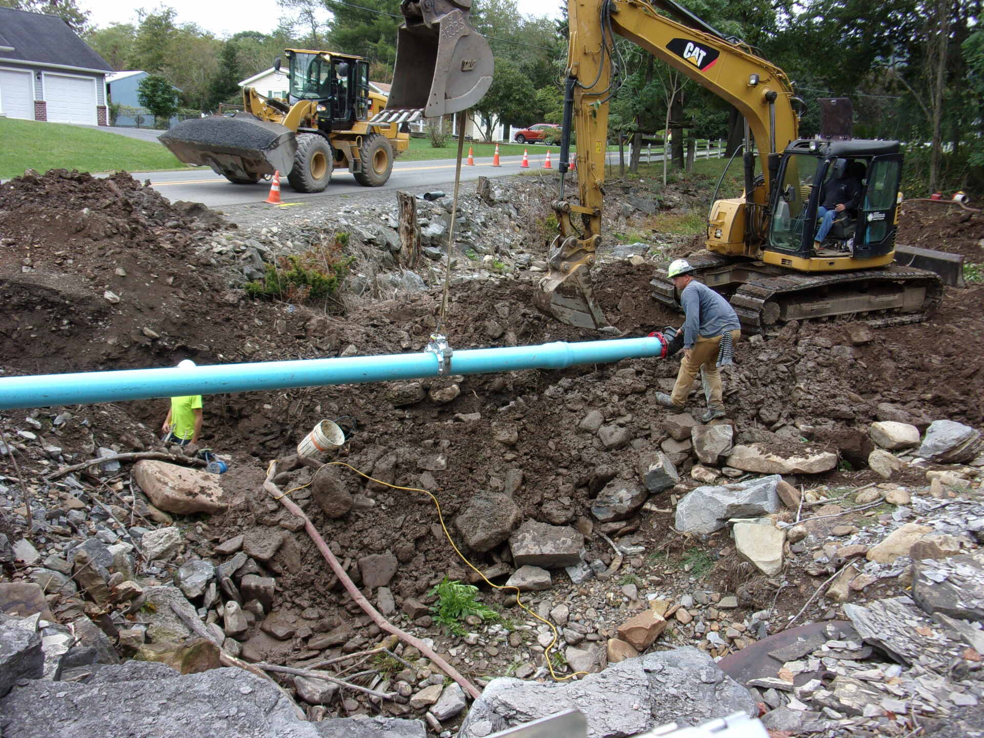 Excavation site with workers and machinery installing a large blue water pipe in a rocky trench, guided by an excavator.
