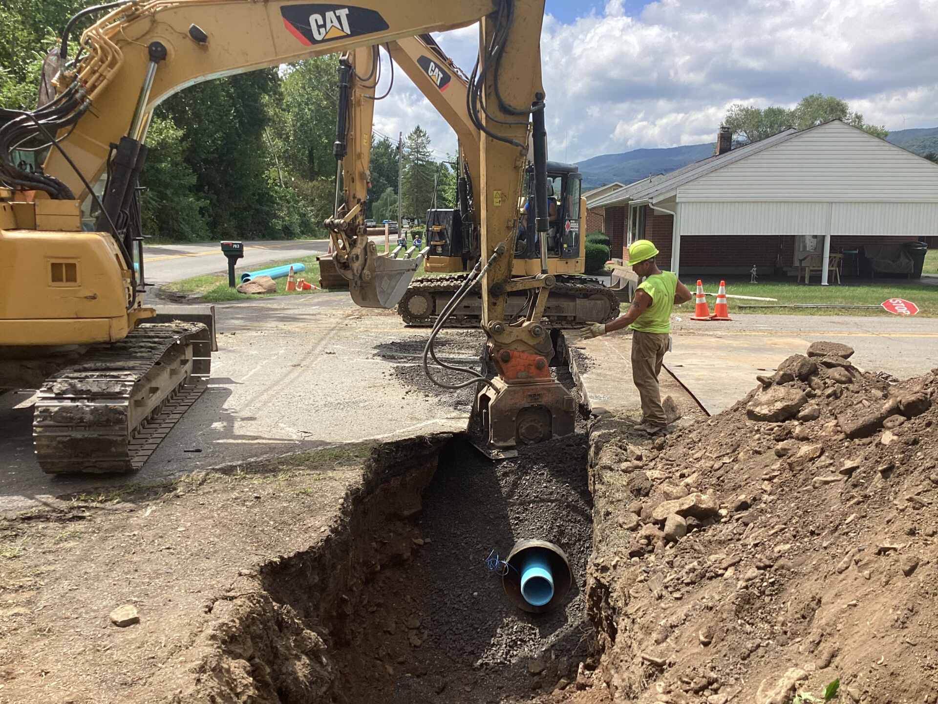 Construction site with an excavator lowering a large black pipe into a deep trench, while workers in safety gear guide the installation.