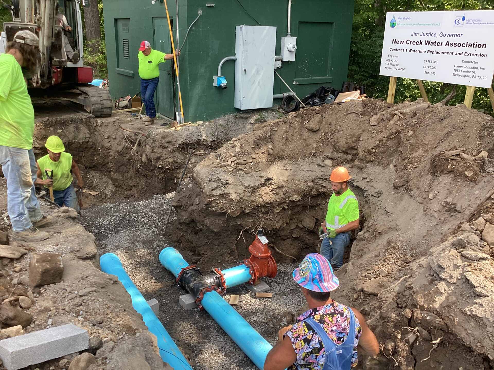Construction site with workers installing large blue water pipes in a deep trench, near a green utility building and a project sign for New Creek Water Association.