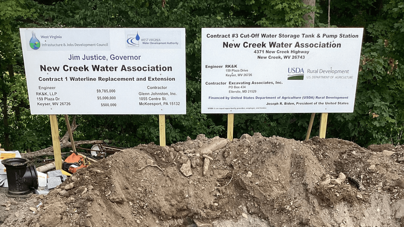 Excavation site with workers and an excavator installing a long blue water pipe in a rocky trench, surrounded by dirt and construction equipment.