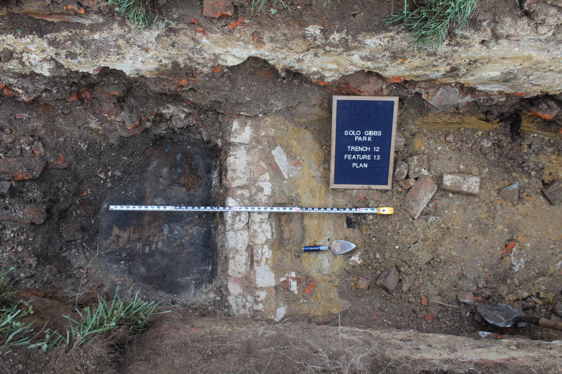 Excavated soil profile showing distinct layers, including a dark charred section and lighter sediment, with a measuring scale and labeled board.