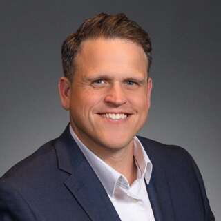 Person in a dark suit jacket and white dress shirt against a plain gray background.