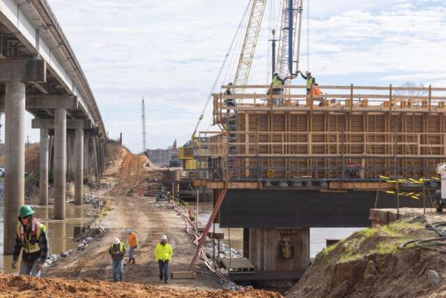 Bridge construction site with workers in safety gear on a large wooden formwork structure supported by steel beams, cranes in the background, and an adjacent roadway with concrete pillars extending over water.