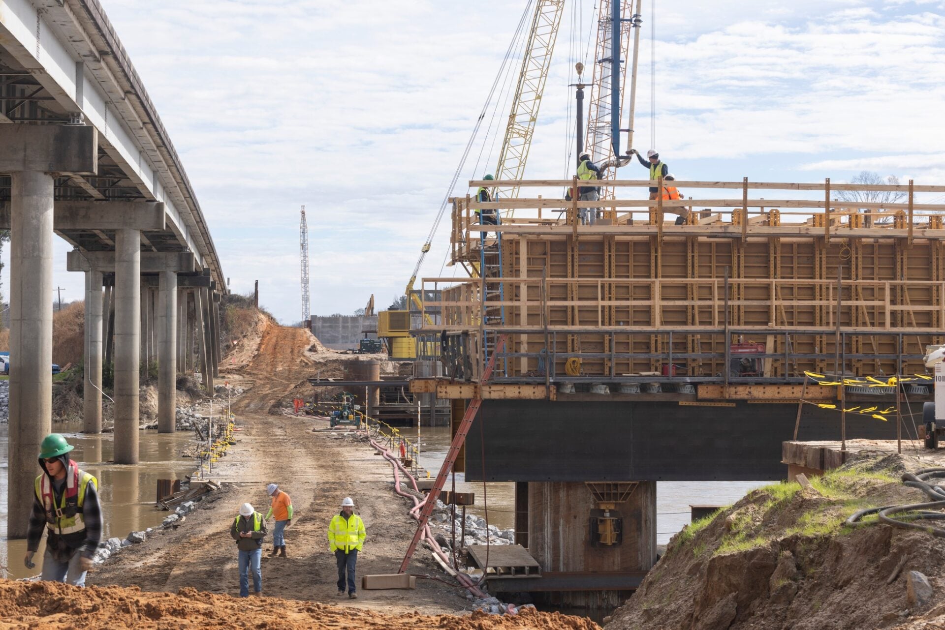 Bridge construction site with workers in safety gear on a large wooden formwork structure supported by steel beams, cranes in the background, and an adjacent roadway with concrete pillars extending over water.