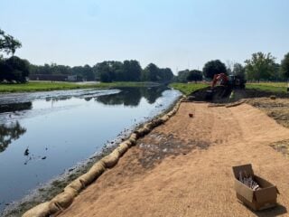 Restoration work along a creek shoreline with erosion control matting, sandbags, and construction equipment near the water.