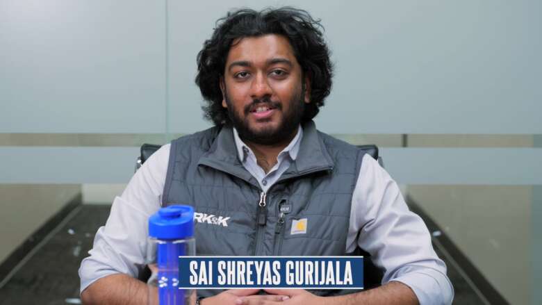 Sai Shreyas Gurijala seated at a conference table with a water bottle, wearing a gray vest in a glass-walled meeting room.