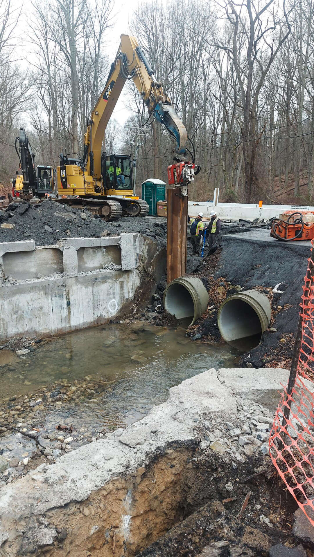 Excavator installs steel sheet piling beside twin culvert pipes in a stream as part of foundation and drainage work for the Henryton Road Bridge Replacement.