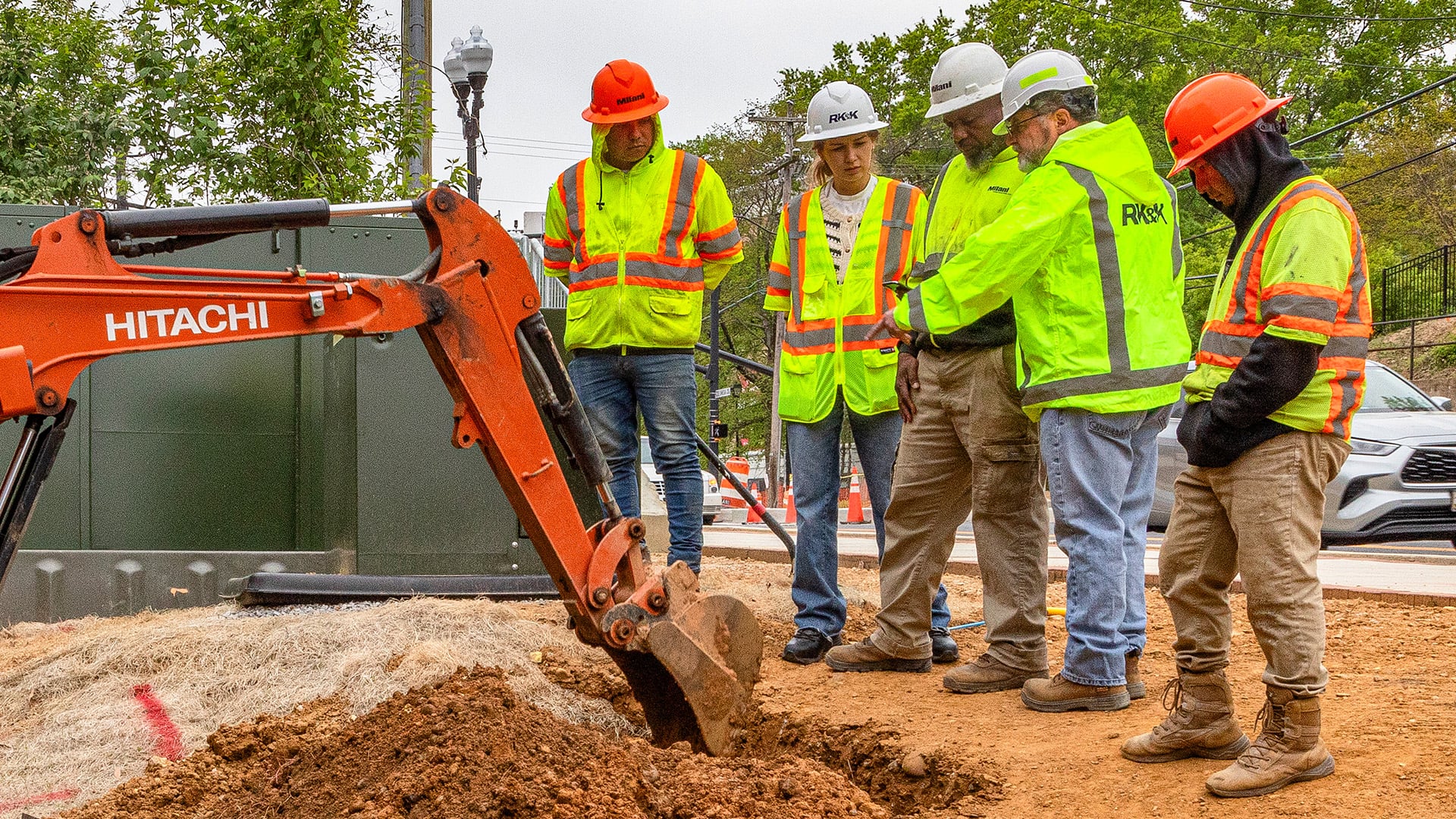 Construction crew in safety gear inspecting a trench beside a Hitachi excavator at a roadwork site.