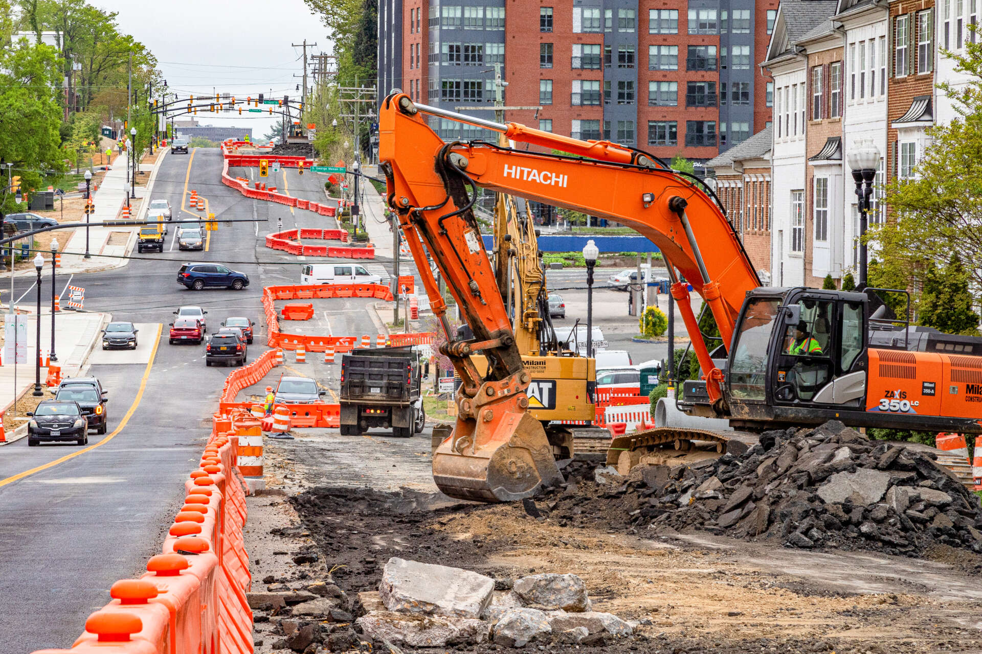 Excavator removing pavement along a busy roadway lined with orange construction barriers and traffic lanes under reconstruction.