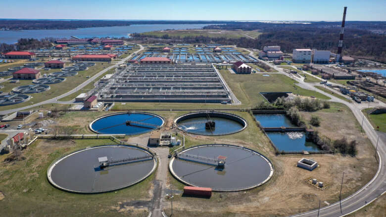 Aerial view of a large water and wastewater treatment facility with circular clarifier tanks, rectangular treatment basins, support buildings, and a nearby reservoir along the shoreline.